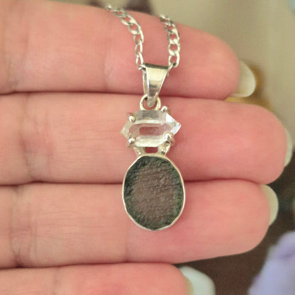 A silver-colored pendant featuring a moldavite and a herkimer diamond, held in a person's hand.