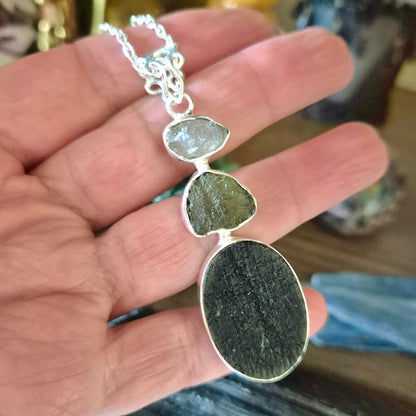 A sterling silver pendant featuring a moldavite teardrop, a herkimer diamond, and a black tourmaline oval, held in a person's hand.