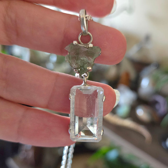 A hand holding a pendant that features an emerald cut Lemurian Seed Crystal above a raw Moldavite, set against a blurred background.