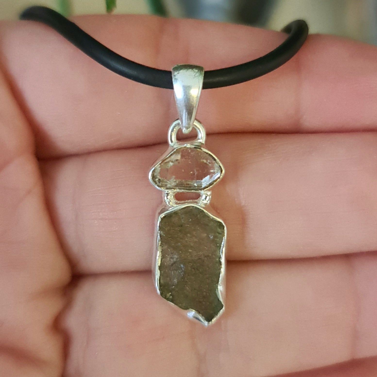 A Moldavite & Herkimer Diamond Pendant with a silver setting, suspended from a black necklace cord, held between fingers.