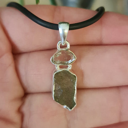A Moldavite & Herkimer Diamond Pendant with a silver setting, suspended from a black necklace cord, held between fingers.