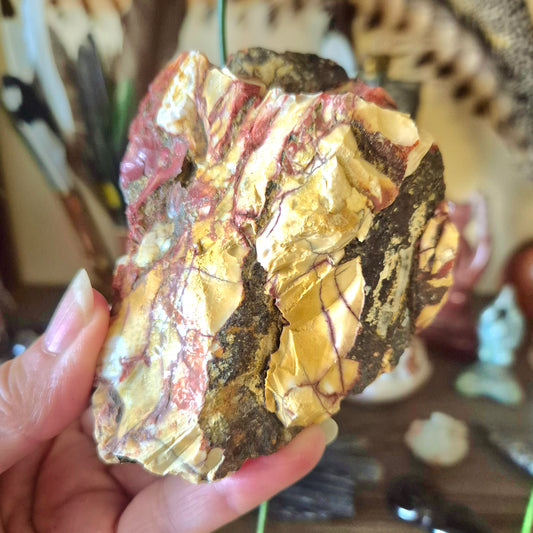 A close-up image of a person's hand holding a Mookaite Jasper Chunk, with a blurred background containing various objects.