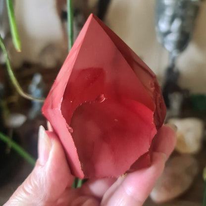 A polished red Mookaite Jasper crystal point held in a person's hand, with a blurred background suggesting indoor setting