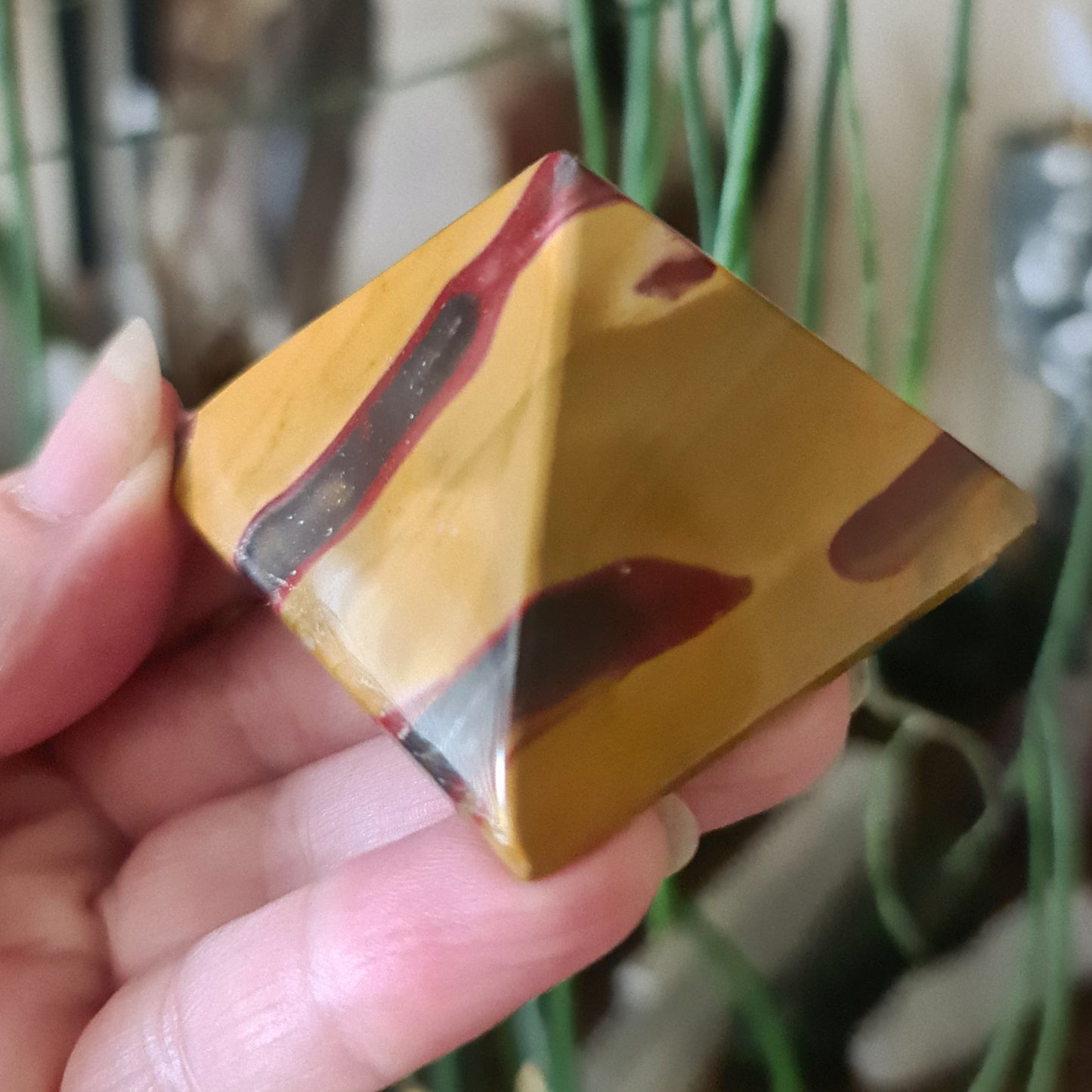 A polished Mookaite Jasper crystal with a geometric pyramid shape, held in a person's hand.