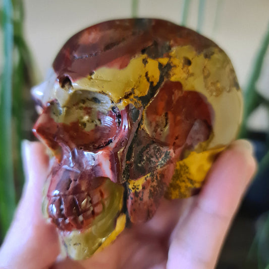 A skull-shaped Mookaite Jasper stone with red, yellow, and brown colors, held in a person's hand.