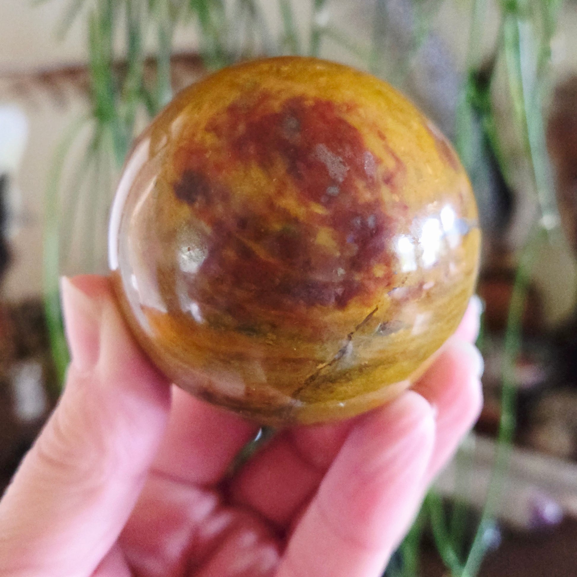 A hand holding a polished Mookaite Jasper sphere with a smooth surface and a camouflage pattern of brown and white colors.