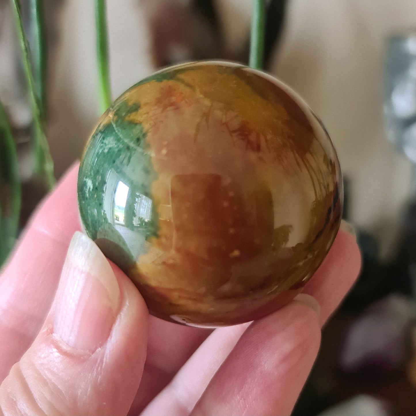 A hand holding a Mookaite Jasper sphere with green and brown colors.