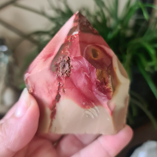 A polished point of Mookaite Jasper held in a person's hand, with a reddish-brown color and natural variations.