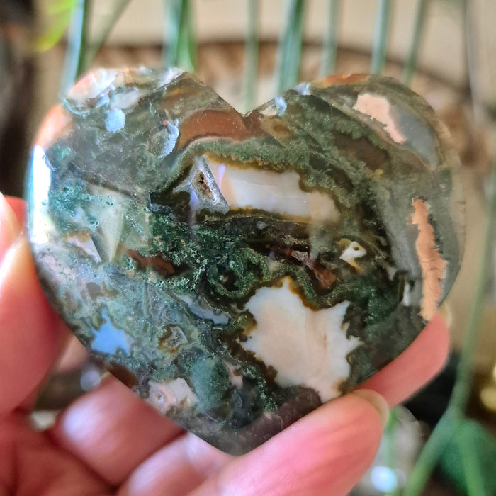 A hand holding a heart-shaped moss agate stone with a blurred background.