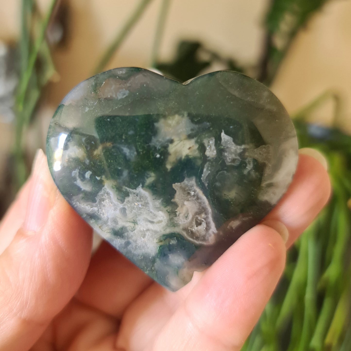 A heart-shaped moss agate stone held in a person's hand, with a green and brown color pattern.