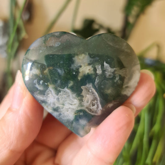 A heart-shaped moss agate stone held in a person's hand, with a green and brown color pattern.
