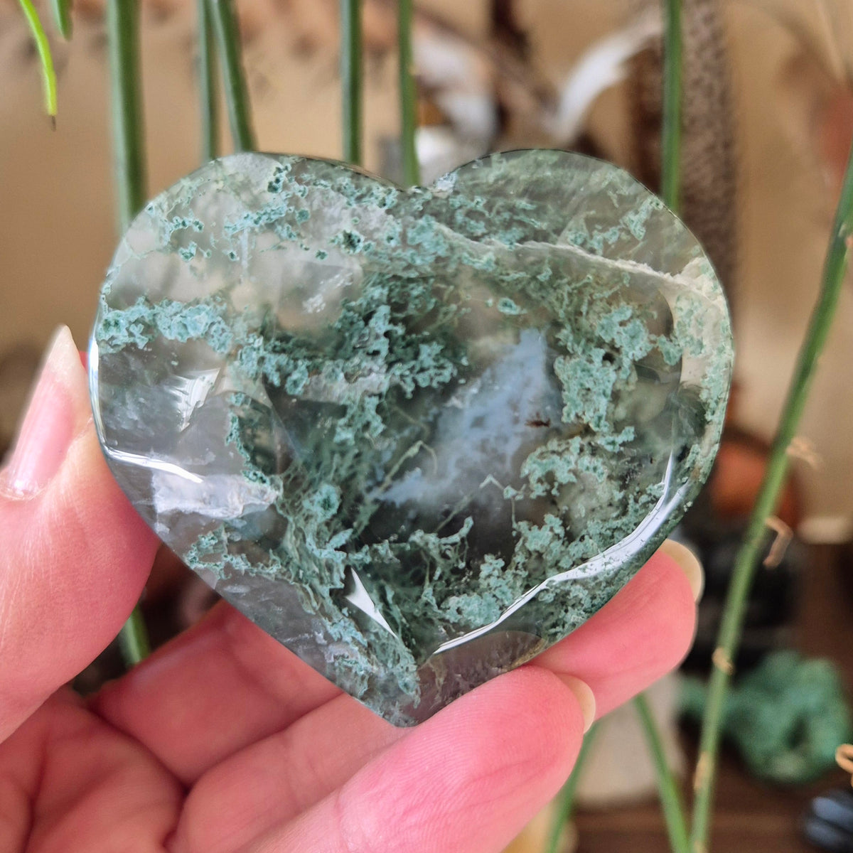 A heart-shaped moss agate stone held in a hand, with a visible green and white layered pattern.