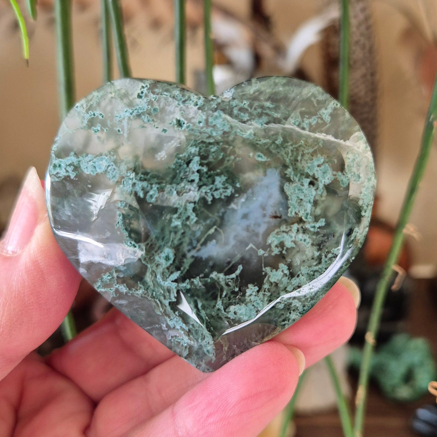 A heart-shaped moss agate stone held in a hand, with a visible green and white layered pattern.