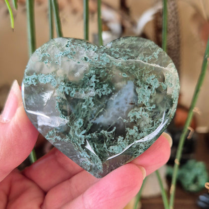 A heart-shaped moss agate stone held in a hand, with a visible green and white layered pattern.