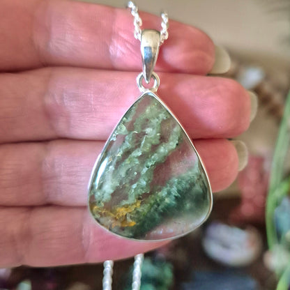 A silver-colored moss agate pendant teardrop shape with a chain attached, held in a hand.