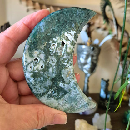 A moon-shaped moss agate stone held in a person's hand, with a blurred background featuring a statue and decorative items.