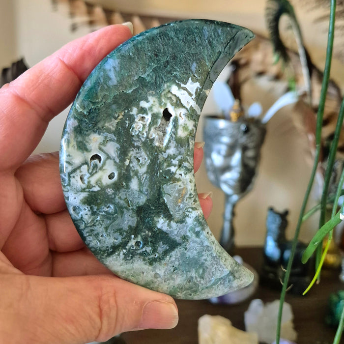 A moon-shaped moss agate stone held in a person's hand, with a blurred background featuring a statue and decorative items.