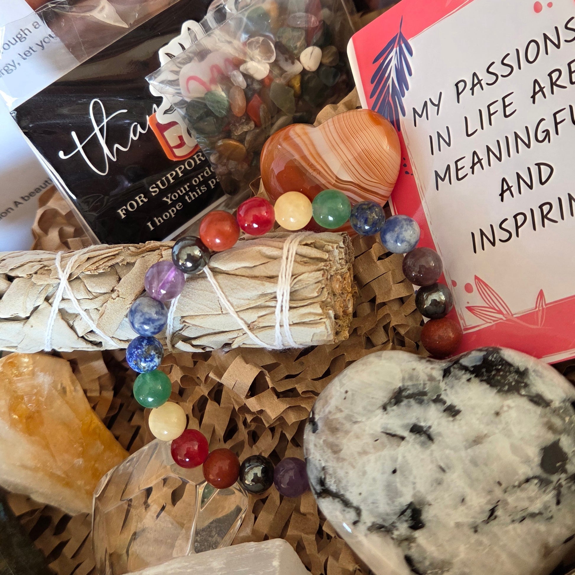 A gift basket filled with crystals and spa items, including a white sage bundle wrapped in string, a beaded bracelet with multicolored round beads, a large polished stone, a yellow crystal, a shell, and a card that reads 'MY PASSIONS IN LIFE ARE MEANINGFUL AND INSPIRING'.