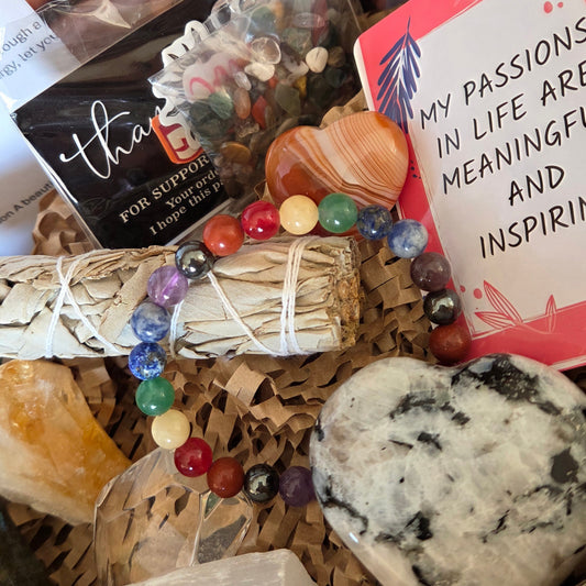 A gift basket filled with crystals and spa items, including a white sage bundle wrapped in string, a beaded bracelet with multicolored round beads, a large polished stone, a yellow crystal, a shell, and a card that reads 'MY PASSIONS IN LIFE ARE MEANINGFUL AND INSPIRING'.