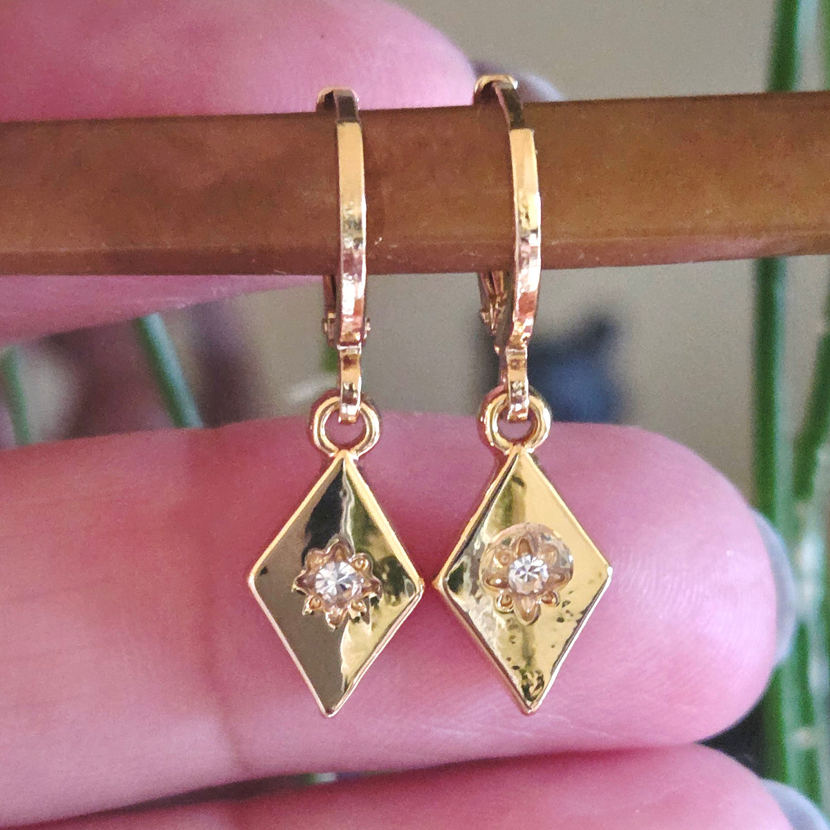Gold diamond-shaped earrings with a central gemstone held between fingers.