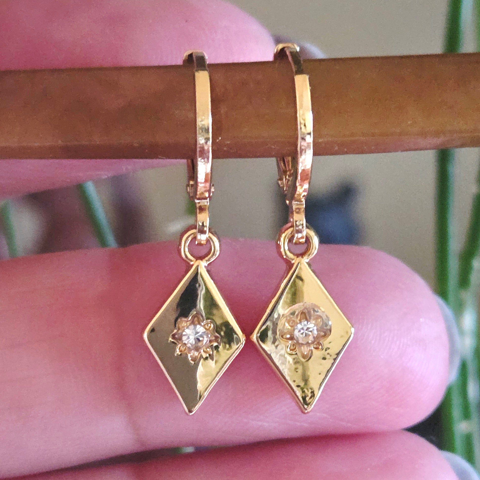 Gold diamond-shaped earrings with a central gemstone held between fingers.