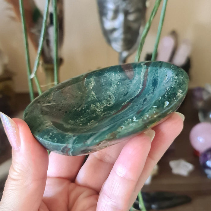 A hand holding a polished green Ocean Jasper stone, with a face and body shaped like a bowl.