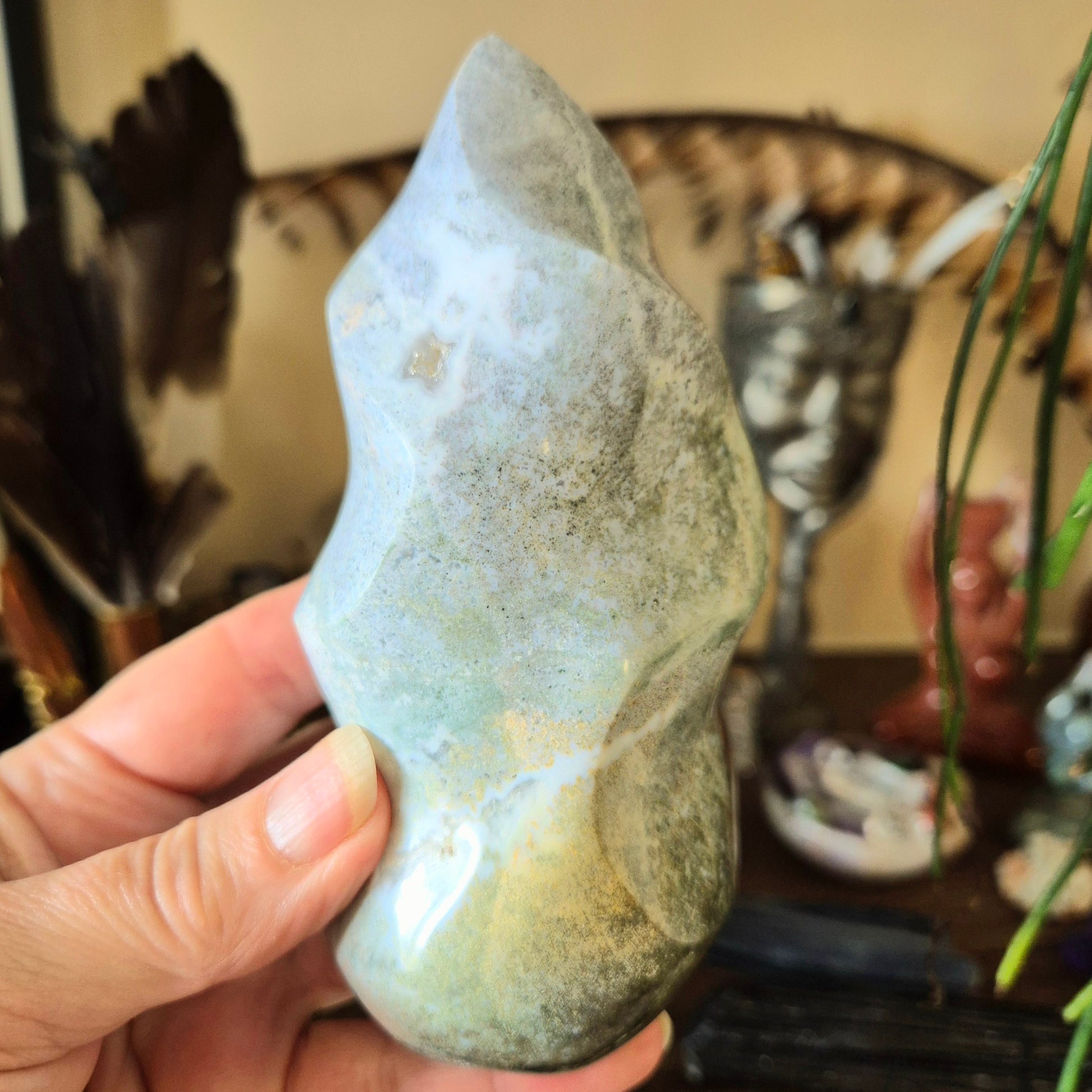 A hand holding an Ocean Jasper gemstone with a flame-like shape, with a blurred background containing various items.