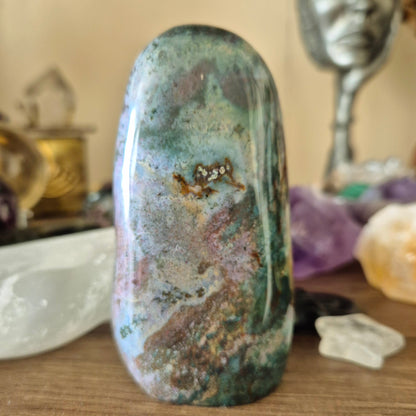 An Ocean Jasper Freeform stone weighing 320 grams, with a green and blue marbled appearance, placed on a wooden surface with crystal and metallic decorations in the background.