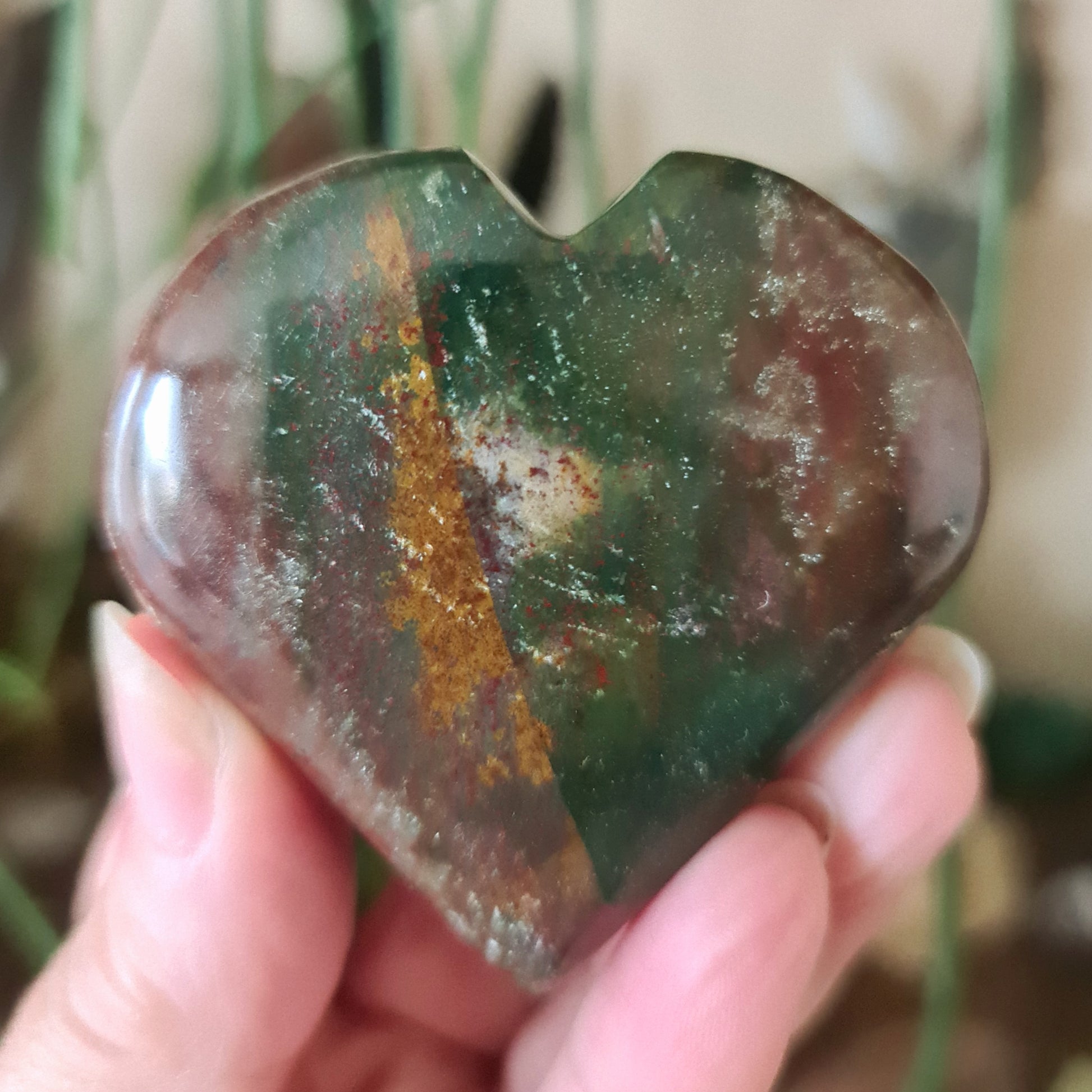 A polished heart-shaped stone with a green and brown color pattern, held in a person's hand.