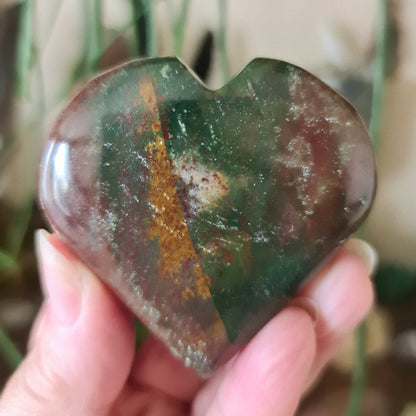 A polished heart-shaped stone with a green and brown color pattern, held in a person's hand.