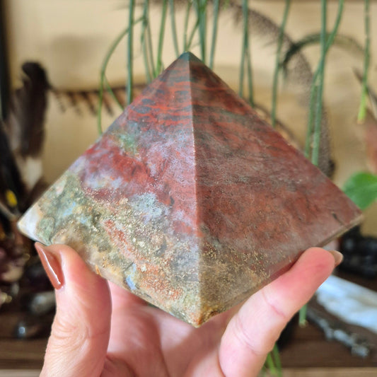 A hand holding a polished jasper stone in a geometric pyramid shape with a marble-like pattern.