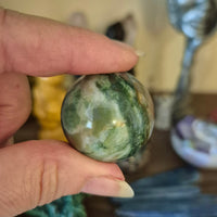 A small Ocean Jasper sphere held in a person's hand, with a blurred background containing various colorful items.