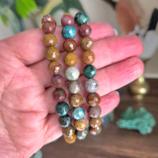 Three beaded bracelets made of faceted gemstone beads in earthy brown, teal, green, gold, and silver tones, held in a person's hand.
