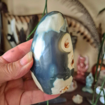 A polished agate freeform sculpture held in a person's hand, with a blurred background featuring various objects and a figure.