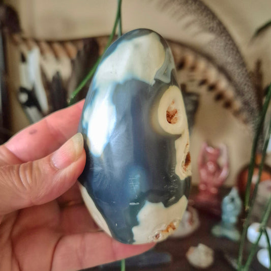 A polished agate freeform sculpture held in a person's hand, with a blurred background featuring various objects and a figure.