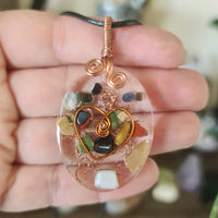 An oval-shaped Orgonite pendant with a mixture of crystals, copper filaments, and wire wrapping, held in a person's hand.