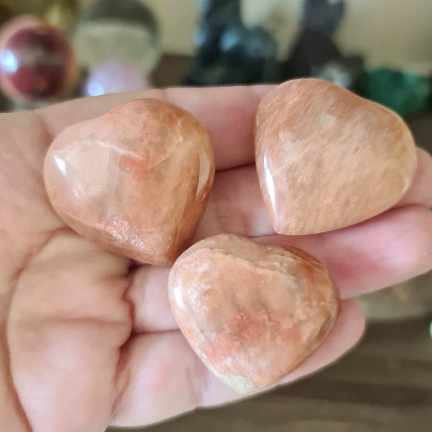 A hand holding three polished Peach Moonstone hearts with a reddish-pink hue.