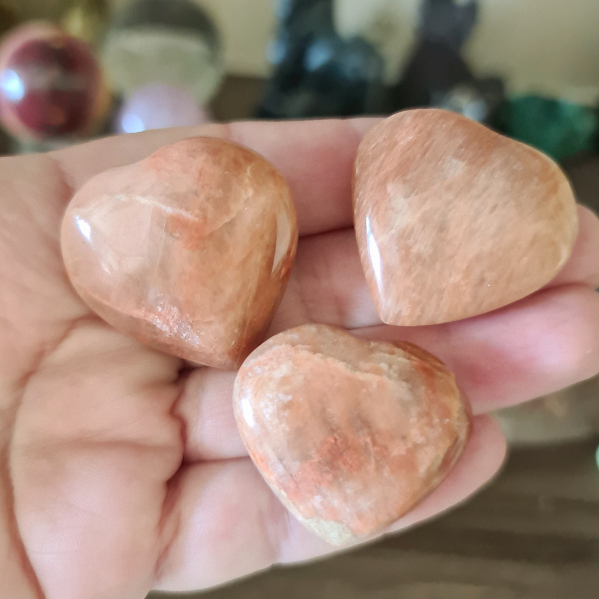 A hand holding three polished Peach Moonstone hearts with a reddish-pink hue.