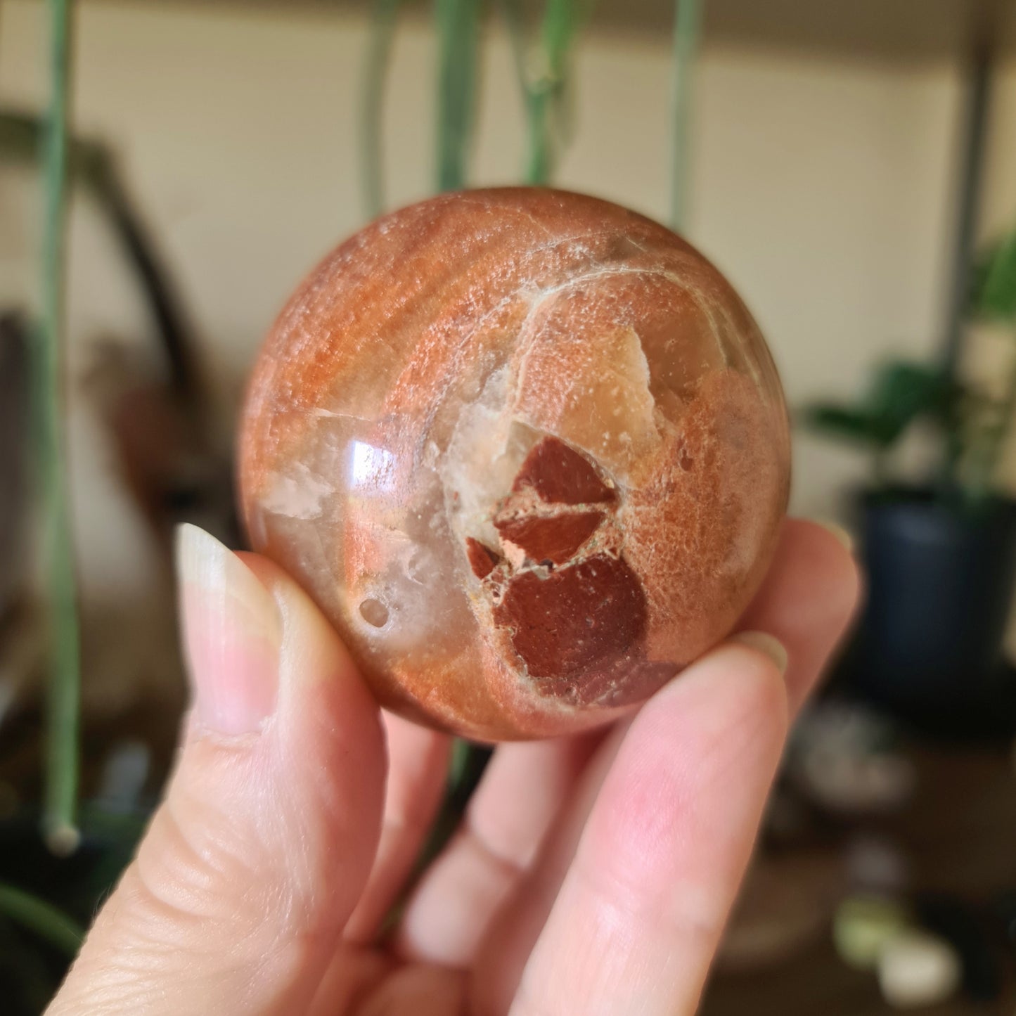 A polished peach moonstone sphere held in a person's hand, with a plant in the background.