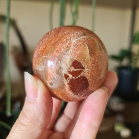A polished peach moonstone sphere held in a person's hand, with a plant in the background.