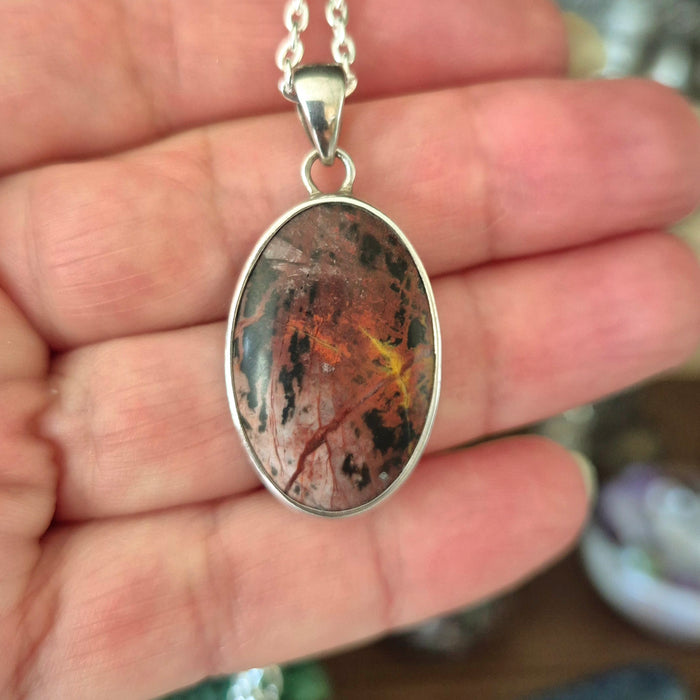 A silver-mounted Pilbara Picasso jasper pendant held in a hand, with a layered red and brown stone design.
