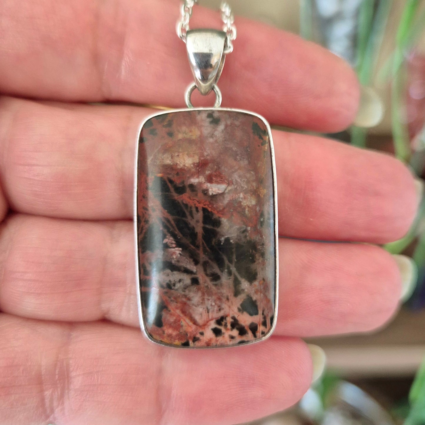A silver-colored jasper pendant with a black and red patterned design, held in a hand.