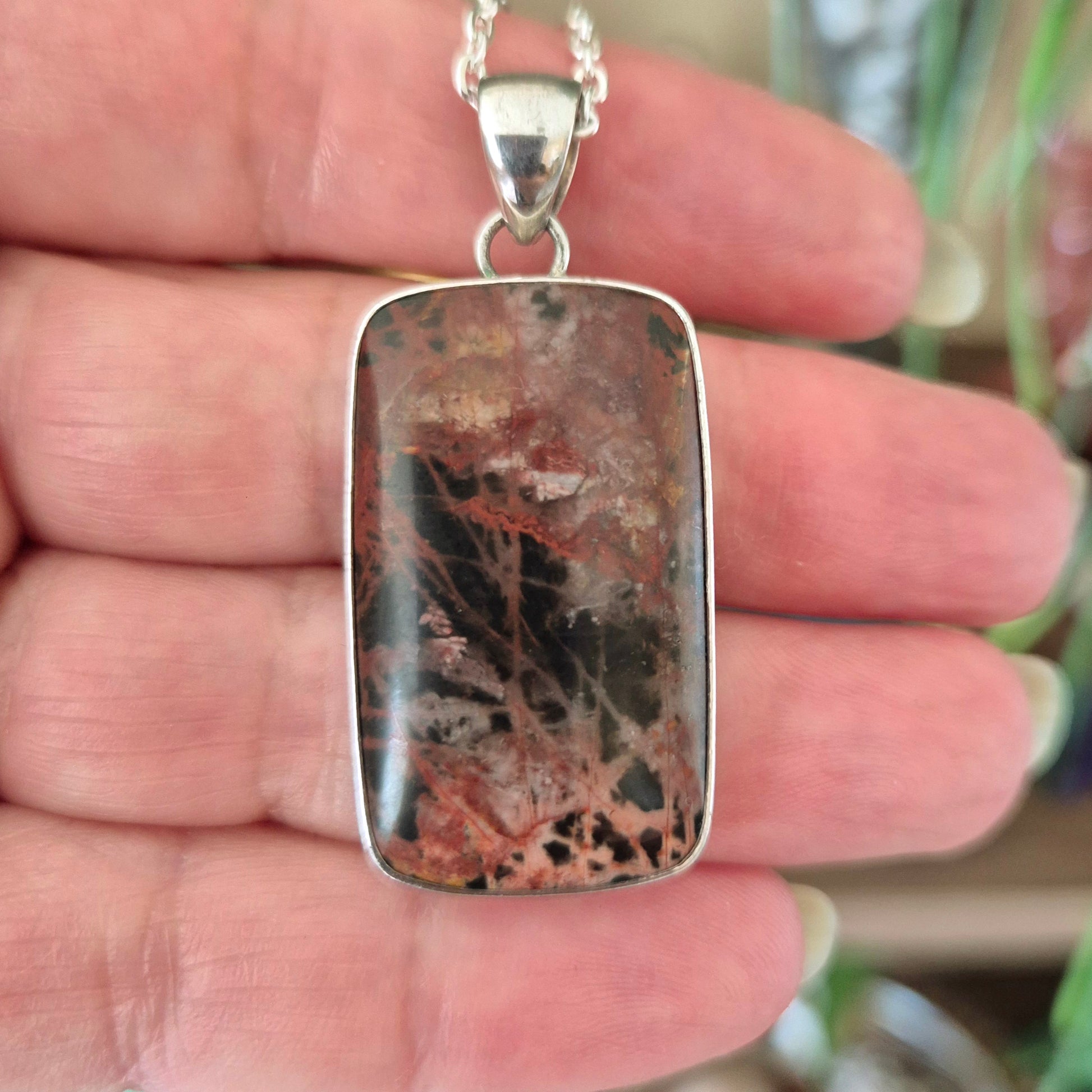 A silver-colored jasper pendant with a black and red patterned design, held in a hand.