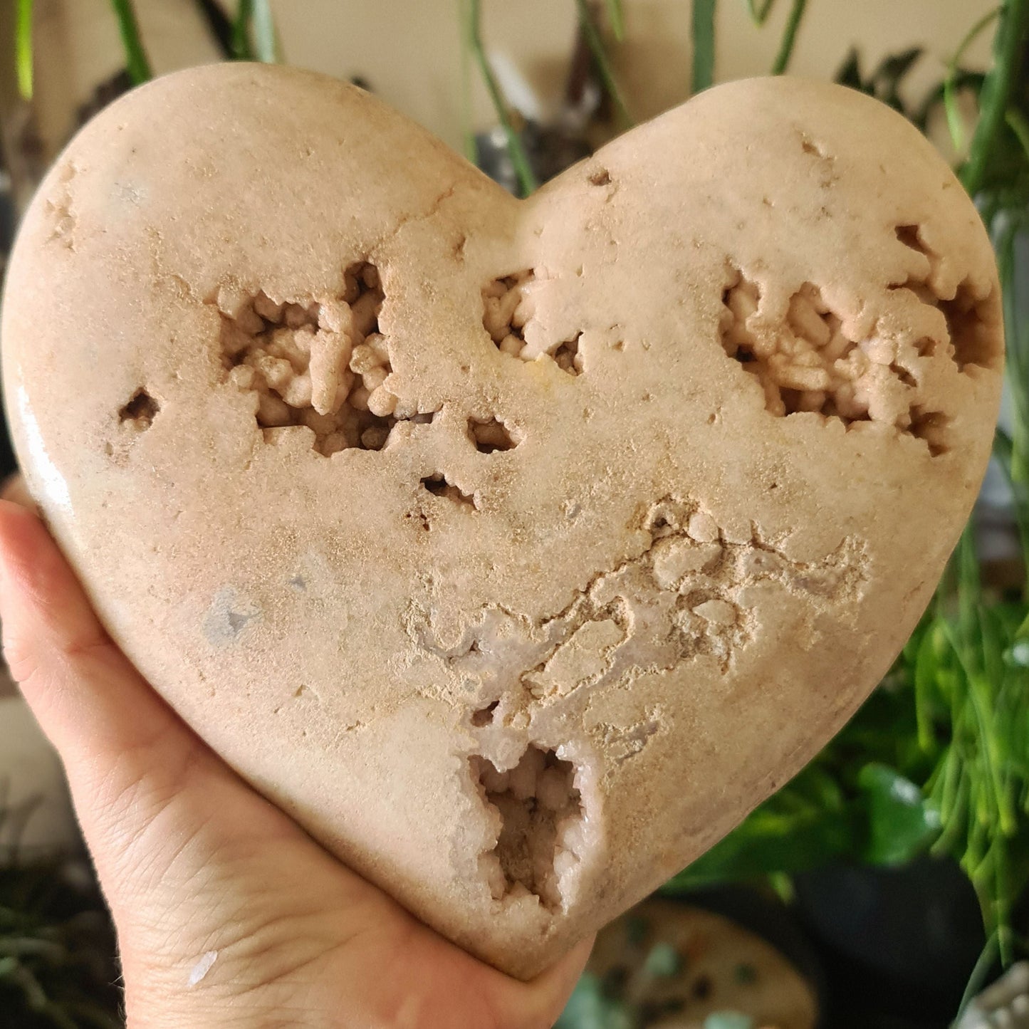 A hand holding a polished pink amethyst heart-shaped crystal with druzy cave inclusions.