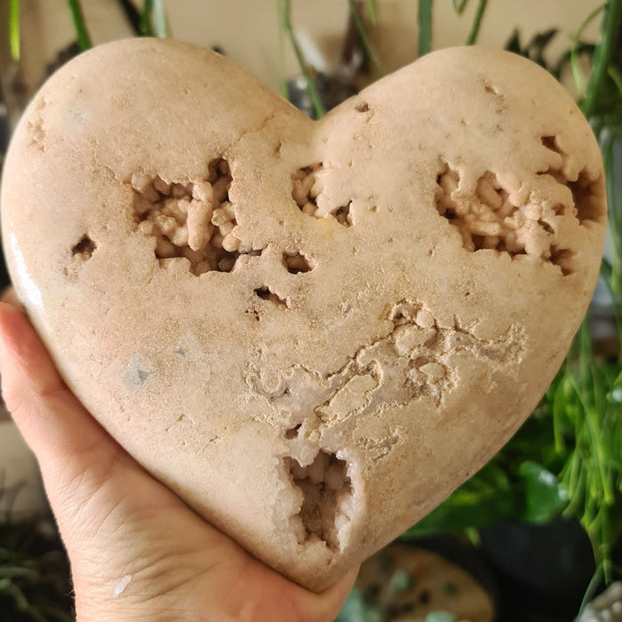 A hand holding a polished pink amethyst heart-shaped crystal with druzy cave inclusions.
