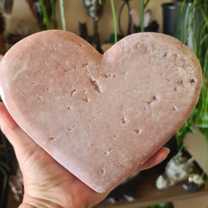 A hand holding a pink amethyst heart-shaped crystal, with a blurred background of various items.
