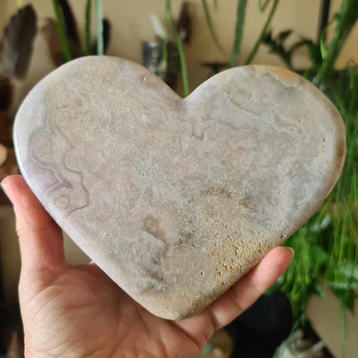 A hand holding a pink amethyst slab heart crystal, with a blurred background of various objects.