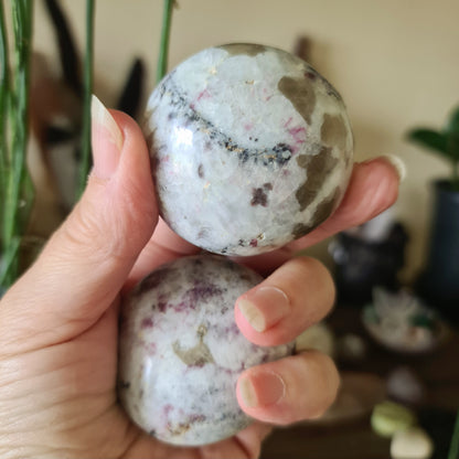 A hand holding a polished pink tourmaline crystal with a palm-like shape, with a background suggesting a natural or outdoor setting.