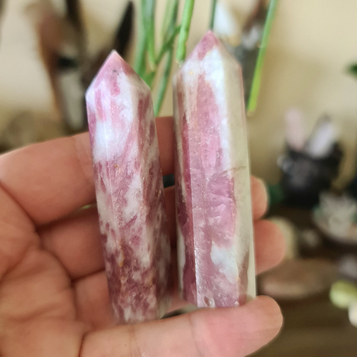 Two polished pink tourmaline crystal points held in a person's hand, with a blurred background suggesting indoor setting