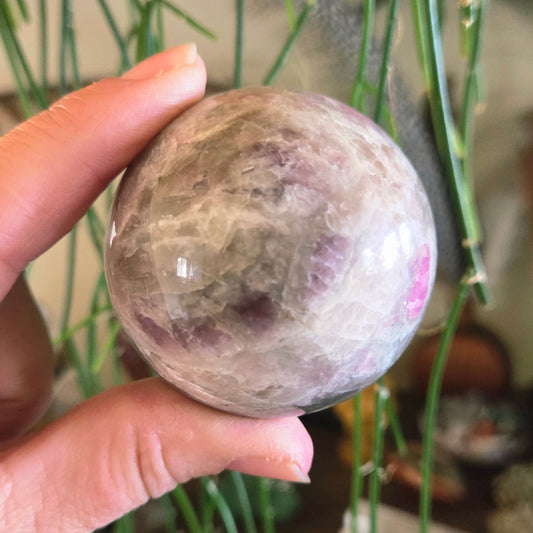 A hand holding a pink tourmaline sphere with a translucent appearance and natural patterns.